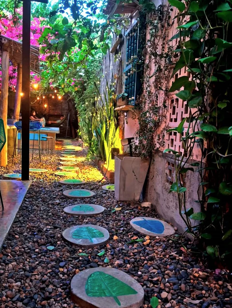 El camino del jardín: piedras redondas con hojas pintadas en verde y azul, luces cálidas y buganvilias al fondo.