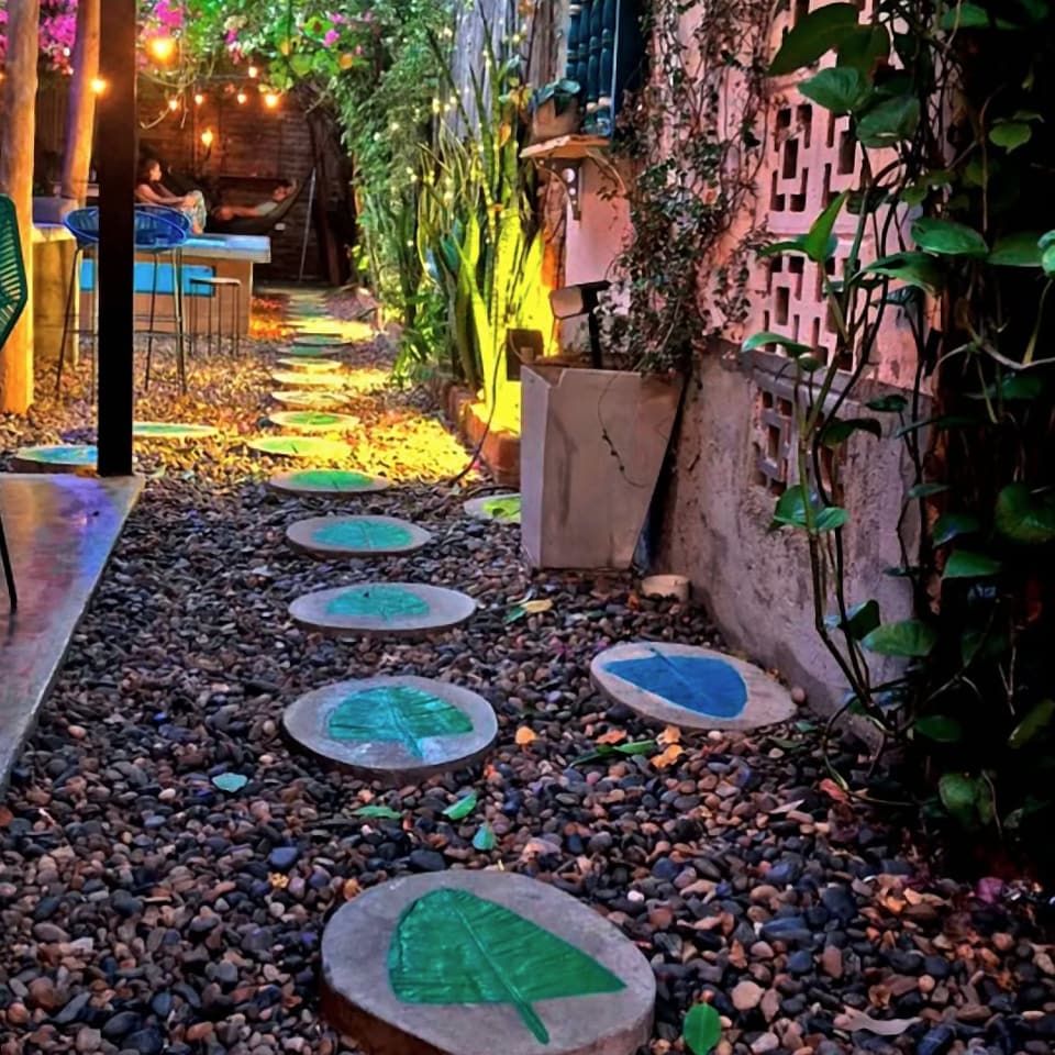 El camino del jardín: piedras redondas con hojas pintadas en verde y azul, luces cálidas y buganvilias al fondo.
