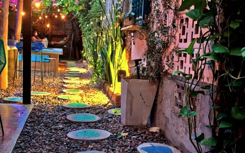El camino del jardín: piedras redondas con hojas pintadas en verde y azul, luces cálidas y buganvilias al fondo.