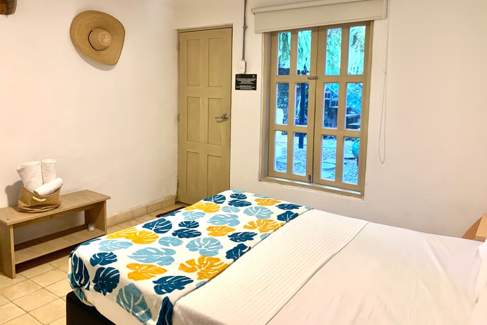 Habitación Doble: cama doble con camino de hojas tropicales en azul y mostaza, ventana y puerta de madera, toallas sobre una banca.
