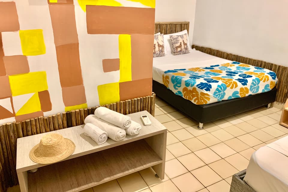 Habitación Triple: mural geométrico en amarillo y terracota, cama doble con hojas tropicales y una banca con toallas y sombrero.